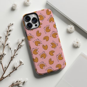 Może przedstawiać: Różowe etui na telefon z wzorem rogalików i napisem "Paris mon Amour" w czarnym skrypcie. Etui ma czarny moduł aparatu. Białe kwiaty i białe tło otaczają etui.
