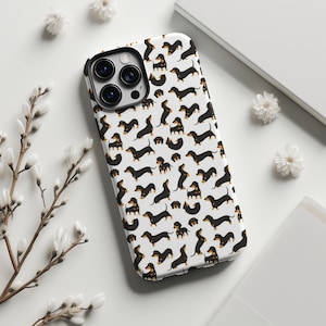 May include: White phone case featuring a repeating pattern of black and tan dachshund illustrations. The case has a black camera lens frame and is surrounded by white flowers and a white background.