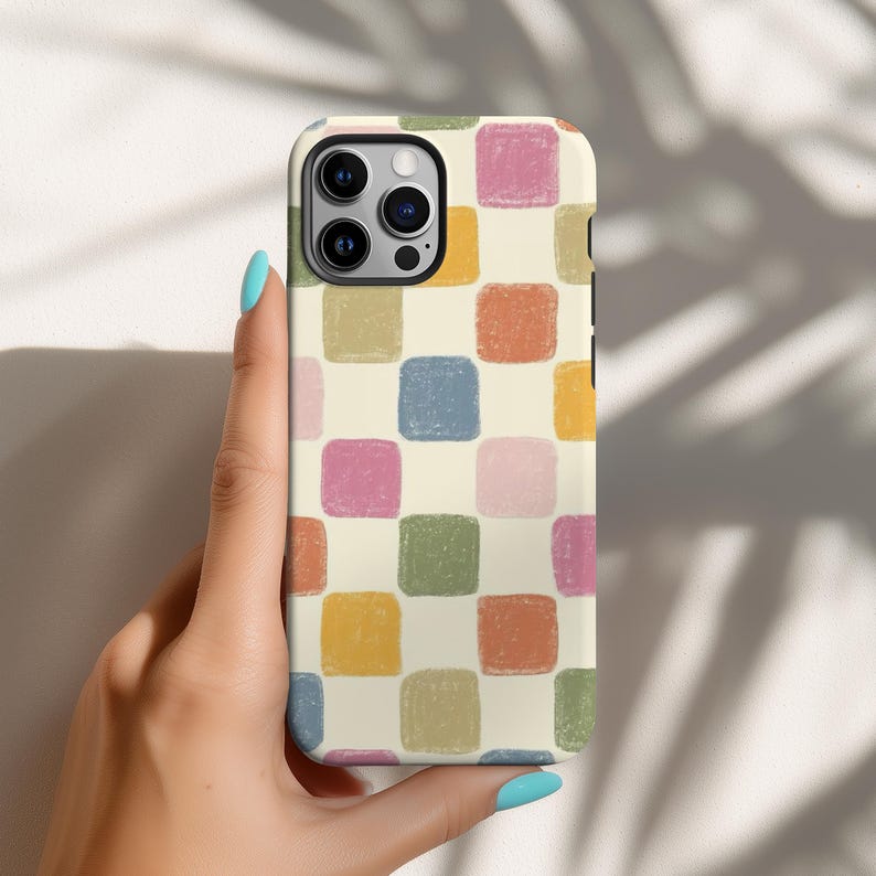 May include: A phone case with a colorful checkerboard pattern. The case features a cream-colored background with squares in shades of pink, blue, green, orange, and yellow. The phone case is held by a hand with blue nail polish.