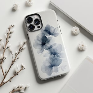 May include: A white phone case featuring a watercolor floral design in shades of blue. The case has a black camera ring and buttons. Delicate white flowers and branches are arranged around the phone case on a white surface.