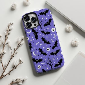May include: A purple phone case featuring a Halloween-themed pattern. The design includes black bats, skulls, bones, stars, and crescent moons. A black camera lens is visible on the phone case.