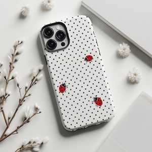 May include: A white iPhone case with a black polka dot pattern and four red ladybug illustrations. The case has a black camera ring and is surrounded by white flowers and a white background.
