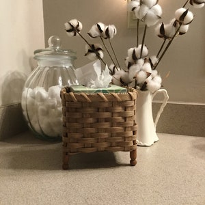 May include: A woven wicker tissue box holder with wooden legs, a clear glass jar filled with cotton balls, and a white pitcher with cotton stems in a white vase.