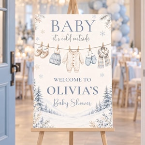 May include: A rectangular welcome sign for a baby shower featuring the text BABY it's cold outside and WELCOME TO OLIVIA'S Baby Shower. The design includes illustrations of baby clothes hanging on a line and a snowy winter forest scene.