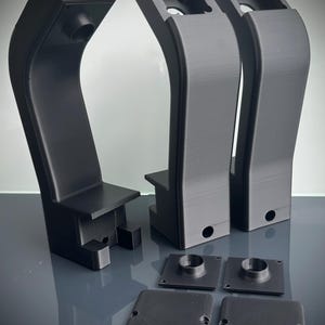 Może przedstawiać: Zestaw czarnych, drukowanych w 3D komponentów. Zestaw zawiera trzy zakrzywione, pionowe konstrukcje z prostokątną podstawą i okrągłym otworem. Obecne są również dwie kwadratowe płyty i dwie okrągłe, cylindryczne części.