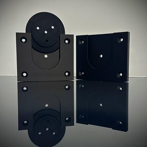 Könnte beinhalten: Vier schwarze Metallhalterungen mit kreisförmigen und rechteckigen Formen. Jede Halterung hat mehrere Löcher zur Befestigung. Die Objekte sind auf einer reflektierenden Oberfläche angeordnet, wodurch ein Spiegeleffekt entsteht. Das Bild ist gut beleuchtet.