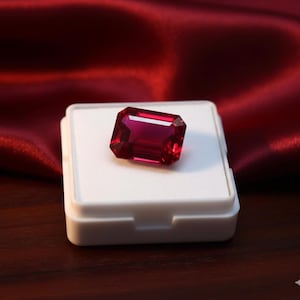 Può includere: Un rubino rettangolare sfaccettato, esposto su una scatola bianca contro uno sfondo di tessuto rosso scuro. Il colore rosso intenso del rubino è accentuato dall'illuminazione, mostrando la sua chiarezza e brillantezza. La gemma è il punto focale.