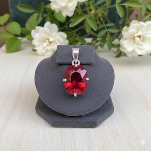 May include: A silver pendant featuring a large, oval-cut, deep red gemstone. The gemstone is set in a silver bezel with small silver accents on each side. The pendant is displayed on a gray jewelry stand against a background of white flowers and green foliage.