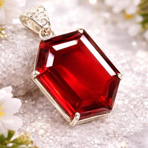 May include: A rectangular, faceted, deep red gemstone pendant with a silver setting. The pendant is displayed on a textured, light grey surface, with white flowers in the background. The gemstone reflects light.