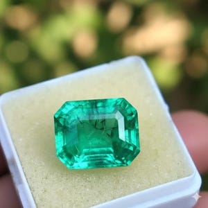 Può includere: Una gemma color verde smeraldo vibrante con taglio smeraldo rettangolare, caratterizzata da inclusioni interne visibili e sfaccettature che riflettono la luce, posizionata all'interno di una piccola scatola espositiva quadrata bianca.