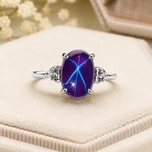 Puede incluir: Un anillo de tono plateado con un gran cabujón de zafiro estrella de color púrpura intenso en forma ovalada, que presenta un patrón de estrella de seis rayos. La piedra central está sujeta por cuatro garras y flanqueada por dos piedras claras.