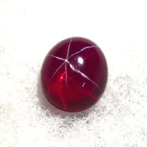 Peut inclure: Une pierre précieuse rouge foncé avec un motif en étoile de lignes blanches rayonnant du centre. La surface polie reflète la lumière, créant un aspect brillant. La gemme est posée sur un fond blanc texturé.