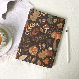 Può includere: Una custodia per tablet marrone scuro con un motivo a tema autunnale. Il design include illustrazioni di foglie, zucche, funghi e altri elementi autunnali in tonalità di marrone, arancione e verde. Uno stilo bianco è posizionato sul lato destro.