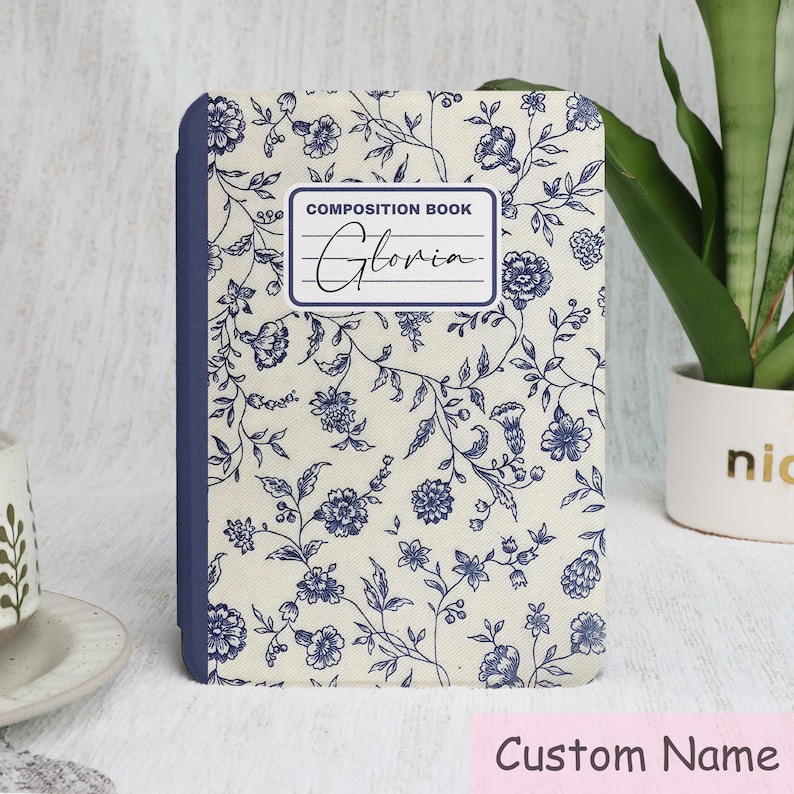May include: A composition book with a floral design in navy blue on an off-white background. The cover features the text "COMPOSITION BOOK" and the name "Gloria" in cursive. The spine is navy blue. The bottom of the image has the text "Custom Name".
