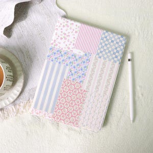 May include: A patchwork tablet case featuring various floral and striped patterns in pastel pink, blue, and white. A white stylus pen rests beside the case. The case is on a white, textured surface.