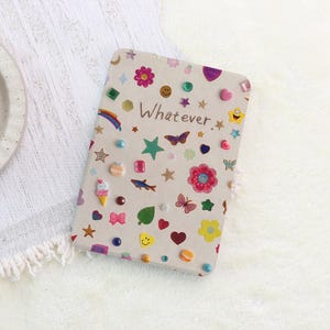 Può includere: Un taccuino rettangolare con una copertina beige chiaro che riporta la parola Whatever stampata al centro. La copertina è decorata con vari adesivi colorati e piccole gemme di plastica, tra cui fiori, stelle, cuori, farfalle e un cono gelato.