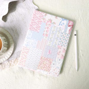 Könnte beinhalten: Eine pastellfarbene Tablet-Hülle mit Patchwork-Design. Die Hülle zeigt verschiedene Stoffe mit Blumen-, Vichy- und anderen Mustern in Rosa, Blau und Weiß. Ein weißer Stift liegt neben der Hülle. Das Design hat eine weiche, feminine Ästhetik mit einer Vielzahl von Mustern.
