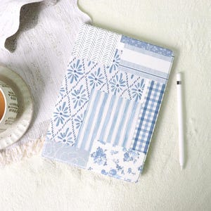 Può includere: Una custodia per tablet in stile patchwork nei toni del blu e del bianco. La custodia presenta vari motivi, tra cui disegni floreali, a righe e a quadri. Una penna stilo bianca è appoggiata accanto alla custodia. L'estetica generale ricorda una trapunta vintage.