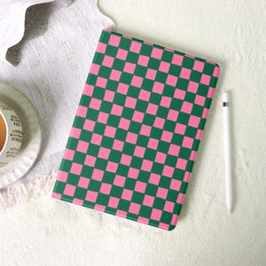 May include: A pink and green checkered tablet case with a white stylus. The case is rectangular and has a repeating pattern of pink and green squares. The stylus is white and has a simple, sleek design. The case and stylus are on a white surface.