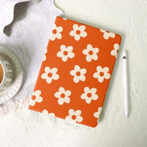May include: An orange tablet case featuring a white daisy pattern. A white stylus is positioned next to the case. The background shows a cup and saucer on a white surface.