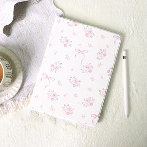 Puede incluir: Una funda rectangular para tableta con fondo blanco y un patrón repetitivo de pequeñas rosas rosadas, hojas verdes y cintas de color rosa claro, acompañada de un lápiz óptico blanco colocado a la derecha de la funda.
