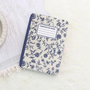 May include: A composition book with a floral pattern in blue and white. The cover features a repeating design of flowers and vines. The spine is a solid blue color, and the cover has a label that reads "COMPOSITION BOOK".