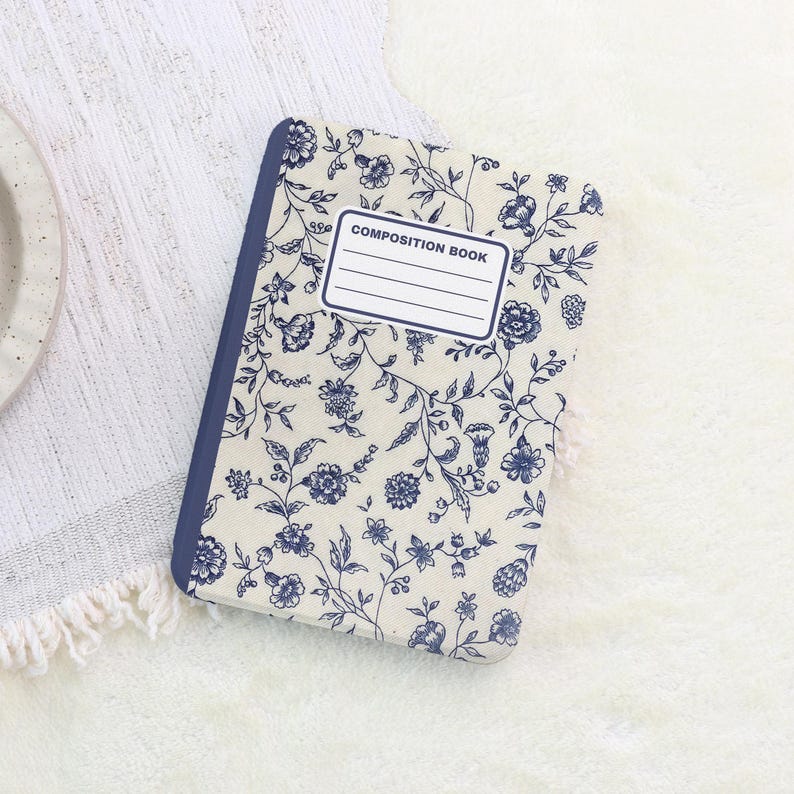 May include: A composition book with a floral pattern in blue and white. The cover features a repeating design of flowers and vines. The spine is a solid blue color, and the cover has a label that reads "COMPOSITION BOOK".