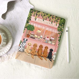 Puede incluir: Una funda rectangular para tableta con una colorida ilustración de siete perros sentados frente al escaparate de una pastelería rosa llena de pasteles. El letrero dice Patisserie y cuelgan enredaderas verdes del borde superior.