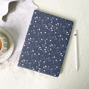 Puede incluir: Una funda de tableta azul marino con un estampado floral de pequeñas flores blancas y azul claro. Un lápiz óptico blanco descansa junto a la funda. La funda está sobre una superficie blanca.