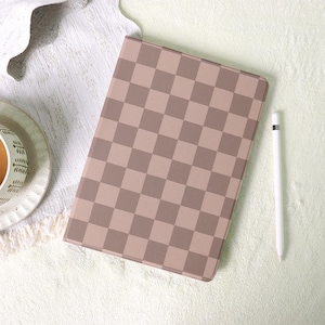 May include: A tablet case featuring a brown and tan chequered design. A white stylus is placed beside the case. The case rests on a white, textured surface, with a cup and saucer in the background.