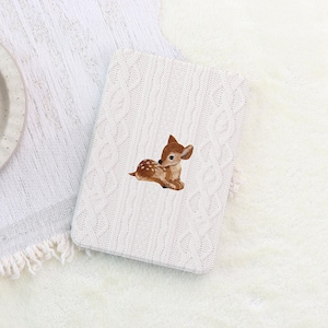 May include: A white, cable-knit tablet case featuring an embroidered fawn. The fawn is brown with white spots and is lying down. The case is rectangular and sits on a white, textured surface.