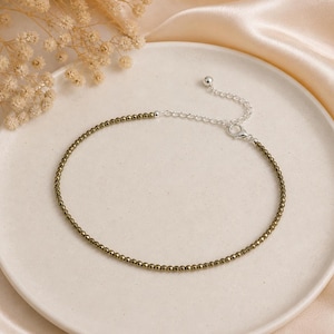 May include: An anklet featuring small, olive-green beads strung together. The anklet has a silver-colored chain and clasp for adjustable sizing. The jewelry is displayed on a white plate with dried flowers in the background.
