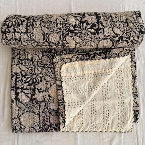 Op de afbeelding: Een opgerolde zwart-beige bloemenquilt. De quilt is gemaakt van een zachte stof en heeft een gedetailleerd botanisch ontwerp. De onderkant is effen crèmekleurig met zwarte stiksels.