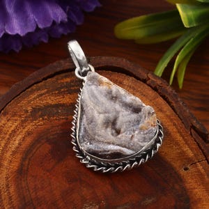 May include: A silver pendant featuring a rough-cut, grey and brown stone. The stone is set in a decorative, twisted silver frame. The pendant has a silver loop for a chain. The background is a wooden surface with green leaves and purple flowers.