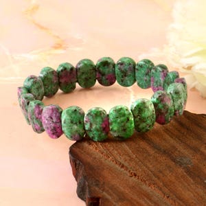May include: A bracelet made of oval-shaped gemstones in shades of green and pink. The beads are strung together to create a circular bracelet. The jewellery is displayed on a wooden surface.