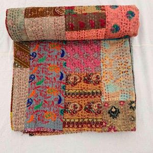 May include: A folded patchwork quilt featuring a variety of rectangular fabric panels in shades of pink, brown, orange, and grey. The textile displays intricate kantha stitching throughout, with diverse floral, paisley, and geometric patterns.