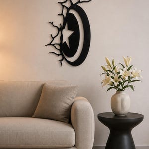 Peut inclure: Une décoration murale en métal noir représentant une étoile dans un croissant, avec un motif en forme d'arbre sur le côté. Elle est fixée sur un mur clair au-dessus d'un canapé beige. Une petite table d'appoint noire supporte un vase blanc avec des fleurs blanches.