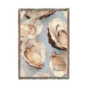 May include: Woven throw blanket featuring a repeating pattern of oyster shells in shades of beige and brown against a light blue background. The blanket has a fringed edge in complementary colors, adding a decorative touch to the design.