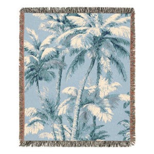 May include: A woven throw blanket featuring a tropical palm tree design in shades of blue and cream. The blanket has a fringed edge in shades of brown and tan. The palm trees are detailed and create a serene, coastal vibe.