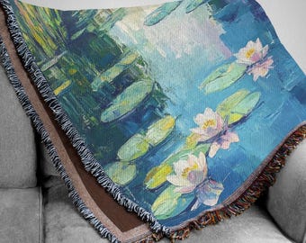 Op de afbeelding: Een geweven sprei met een impressionistisch schilderij van waterlelies in een vijver. Het kunstwerk gebruikt tinten blauw, groen en wit, met gefranjerde randen in verschillende kleuren. De deken is over een grijze bank gedrapeerd.