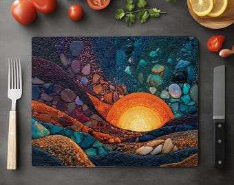 May include: A rectangular glass cutting board featuring a mosaic design of a sunset over a landscape. The artwork uses a variety of colors, including orange, blue, and brown, to depict the sun, sky, and terrain. The board is surrounded by fresh produce.