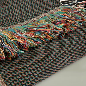 May include: A woven textile with a dark background and a colorful fringe. The fabric has a textured pattern with small, multi-colored dots. The fringe is made of various colored threads, including red, blue, yellow, and green.