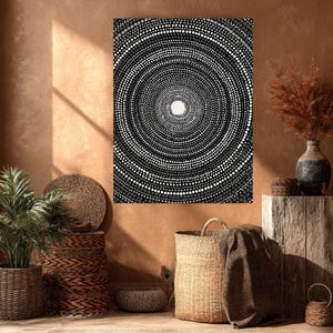May include: Abstract art print featuring a concentric circle design composed of varying sizes of black, white, and gray dots. The artwork is displayed on a wall with a warm, earthy tone, complemented by woven baskets and a plant.