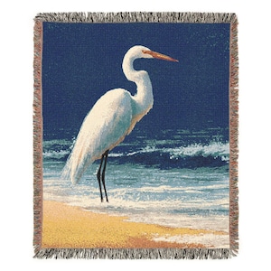May include: Woven throw blanket featuring a white egret standing on a sandy beach with ocean waves in the background. The bird has a long, curved neck and a long orange beak. The blanket has a fringed border and a blue, yellow, and white color scheme.