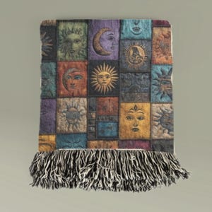 May include: A decorative throw blanket featuring a patchwork design with celestial imagery. The squares depict suns, moons, and faces in various colors, including gold, blue, and purple. The blanket has a black and white fringe along the bottom edge.