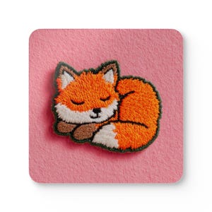 May include: Embroidered patch of a sleeping fox. The fox has orange fur, white markings, and a brown nose and paws. The patch is set on a pink background. The fox is curled up, with its eyes closed, giving a peaceful appearance.