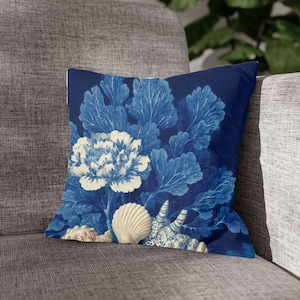 Może przedstawiać: Kwadratowa poduszka dekoracyjna z granatowym tłem i kwiatowo-muszlowym wzorem. Wzór przedstawia duży biało-kremowy kwiat piwonii, niebieskie liście i muszle. Poduszka leży na szarej sofie.