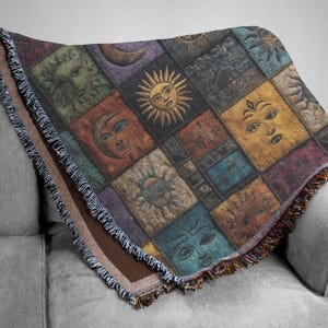 May include: A woven throw blanket featuring a patchwork design with celestial imagery. The squares depict suns, moons, and faces in shades of gold, blue, green, and purple. The blanket has a fringed edge and is draped over a gray sofa.