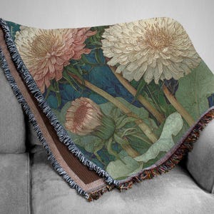 May include: Woven throw blanket featuring a detailed floral design of dandelions in shades of pink, cream, and green against a teal background. The blanket has a fringed edge with brown and tan trim, perfect for adding a touch of nature-inspired decor to any living space.