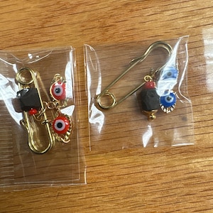 May include: Two gold-colored safety pins with decorative charms. One pin has a black charm and red eye charms. The other pin has a black charm and blue eye charms. The pins are in clear plastic packaging.